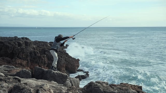 Las olas salpican las rocas mientras un pescador intenta pescar
