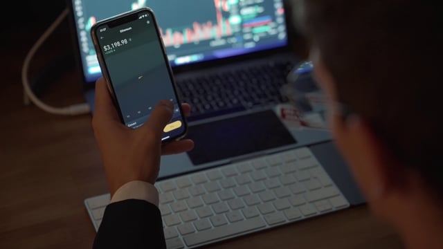 A trader performing financial analysis of cryptocurrency using a phone app and laptop.